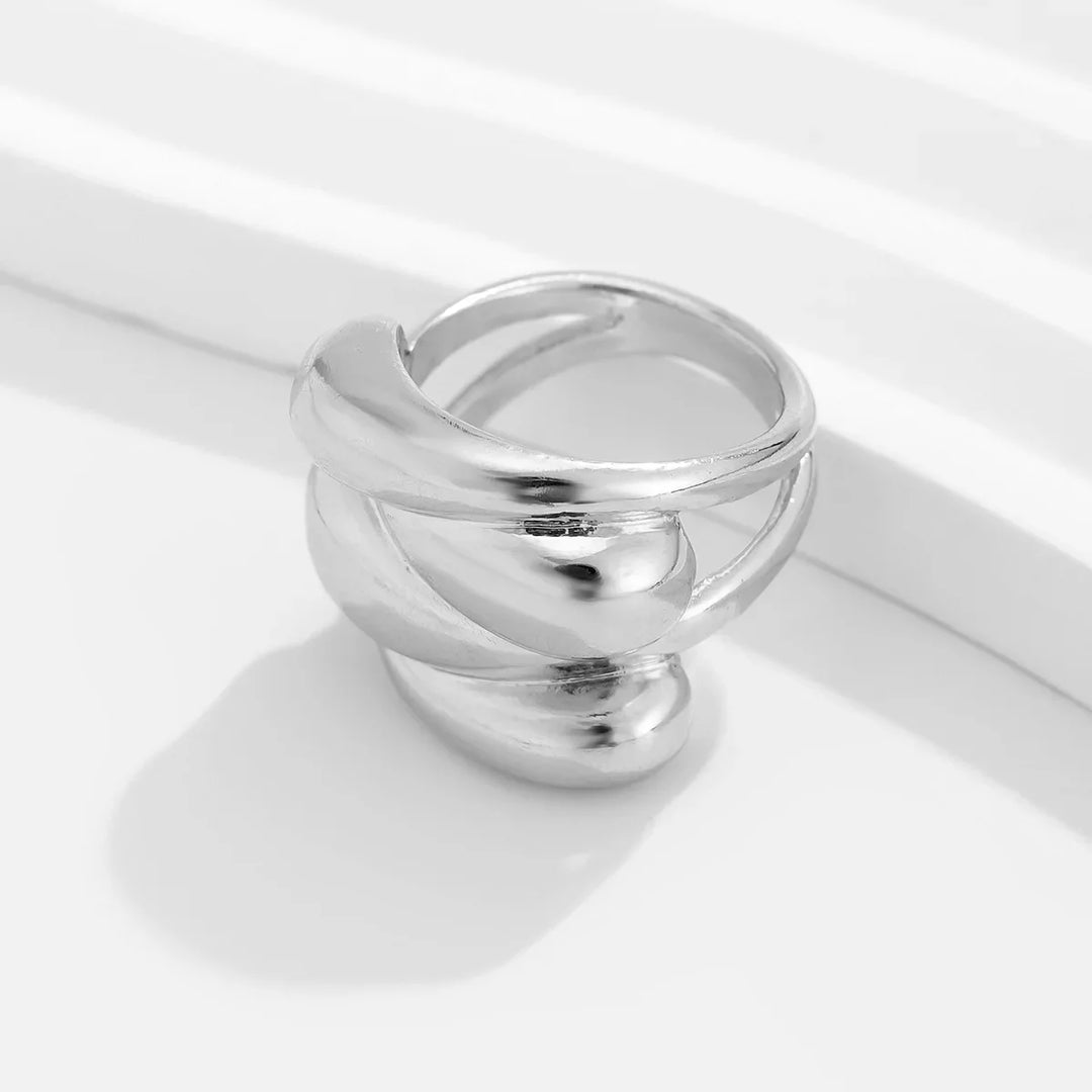 A chunky silver ring with a modern, overlapping wave geometric design is displayed on a smooth white surface with curved lines in the background.
