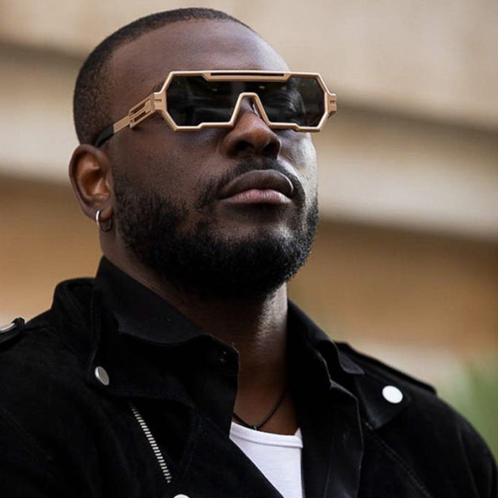 A man with a beard wears bold, heavy metal sunglasses with thick gold square frames and a black jacket, looking confidently into the distance.