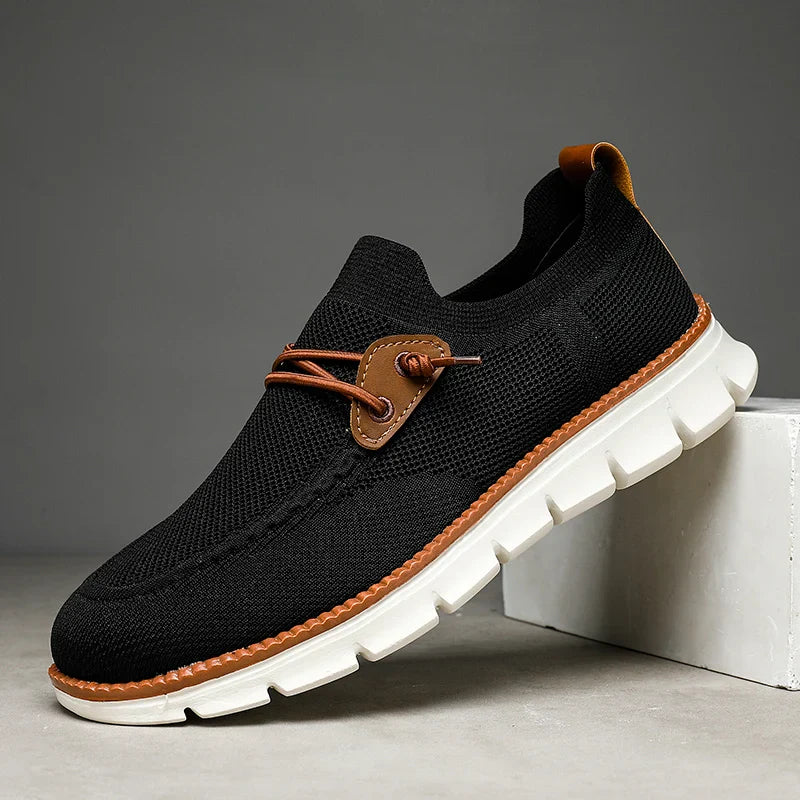 A black slip-on sneaker with a white chunky sole, brown leather lace detail on the side, and a textured knit upper—these breathable loafers are positioned against a neutral background.
