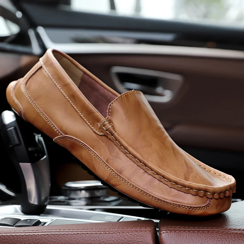 A tan hand-stitched moccasin loafer with visible stitching rests on the center console inside a car, with the cars interior and gear shift in the background.
