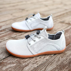 A pair of light gray casual sneakers with breathable mesh uppers, a wide toe box, gray laces, and brown cushioned soles are placed on a wooden surface. One shoe is in the foreground, the other slightly out of focus in the background.