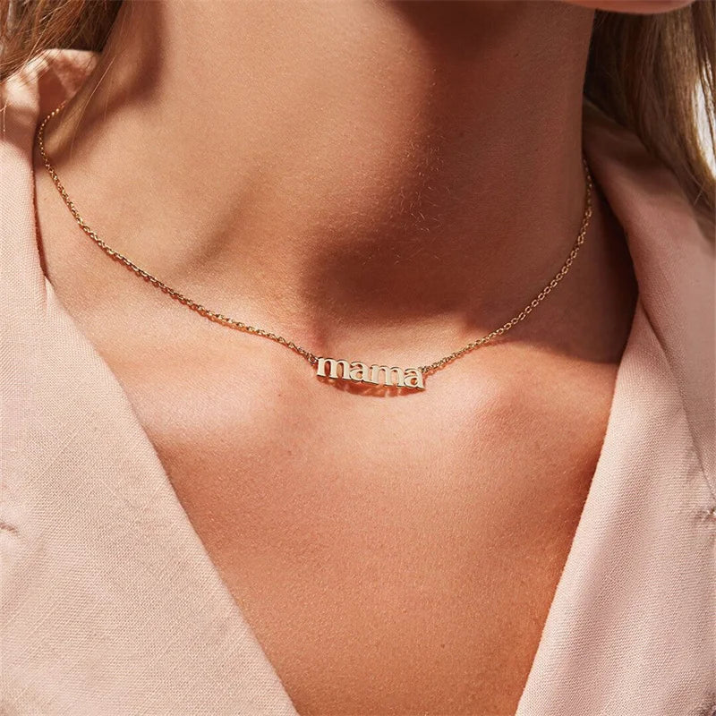 A woman wearing a gold mama pendant necklace in lowercase letters, shown against her neck and light pink blouse—a perfect Mothers Day necklace.