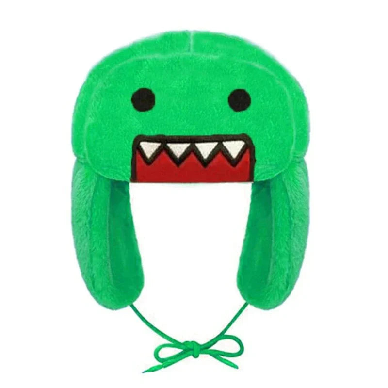 A green plush winter hat with adjustable ear flaps, designed to look like a cartoon monster face with black eyes, white pointy teeth, and a red mouth. The hat has strings for tying under the chin for cozy winter styling.