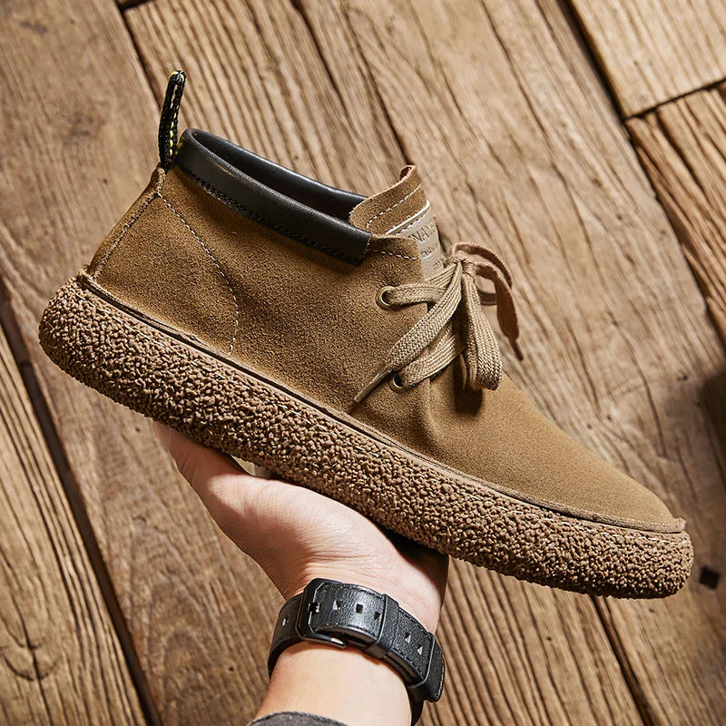 A hand holds a tan cow suede leather casual shoe with brown laces, a textured rubber sole, and a black collar, against a background of wooden floorboards—a stylish pick inspired by classic Oxfords dress shoes.