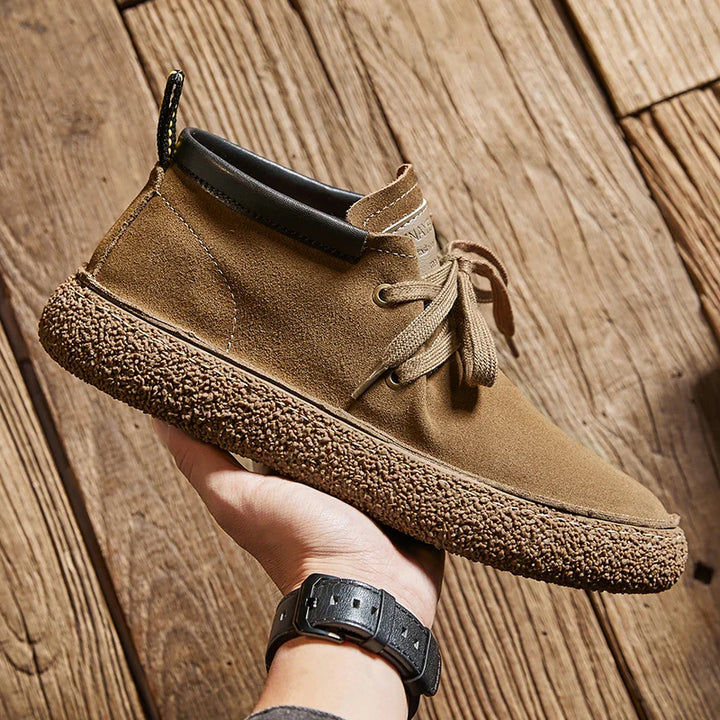A hand holds a tan cow suede leather casual shoe with brown laces, a textured rubber sole, and a black collar, against a background of wooden floorboards—a stylish pick inspired by classic Oxfords dress shoes.