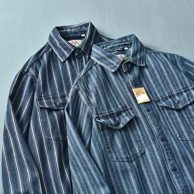 Two blue denim vintage button-up shirts with white vertical stripes are laid flat side by side on a gray surface. Both feature relaxed fit, collars, chest pockets, and front buttons. One shirt has a tag attached to the collar.