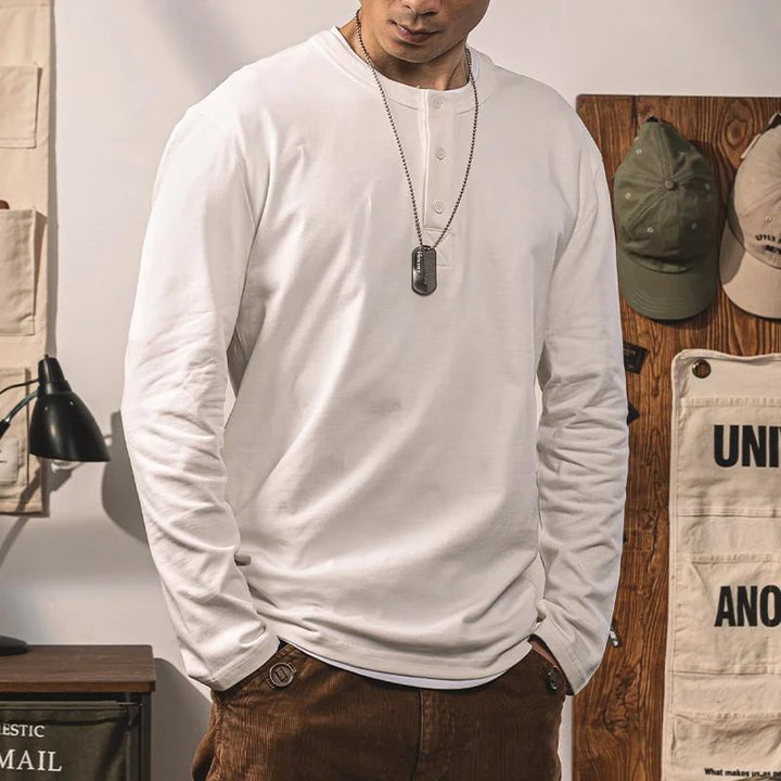 A person wears a pure cotton, long-sleeve Henley shirt, brown pants, and dog tag necklace indoors. The background shows hats on a wooden rack, a wall lamp, and canvas bags. The persons face is partially visible.