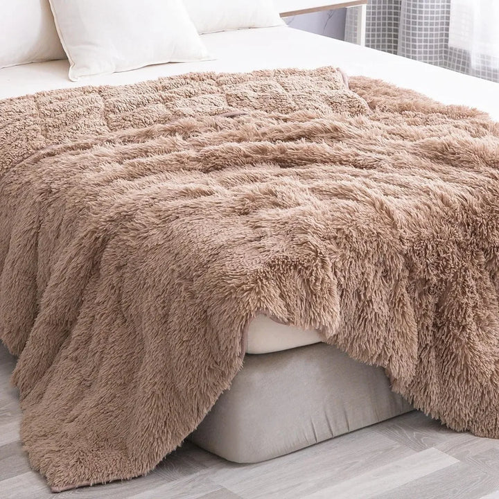 A soft, fluffy Faux Fur Weighted Blanket is draped over the edge of a neatly made bed with white pillows in a bright, modern bedroom with light gray flooring.