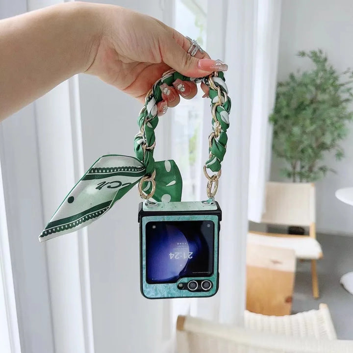 A hand holds a foldable smartphone with a green marble-patterned case, featuring a slim design and a braided green-and-white chain strap decorated with a matching scarf. The background reveals a bright, modern room with plants and light-colored furniture.