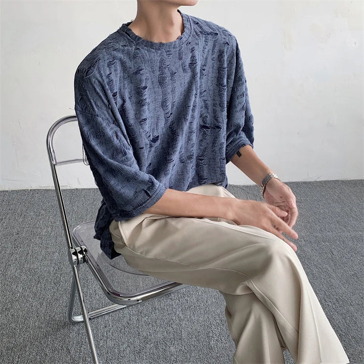 A person in a textured blue oversized tee and beige pants sits sideways on a transparent folding chair against a plain wall and carpeted floor. Their face is not visible.