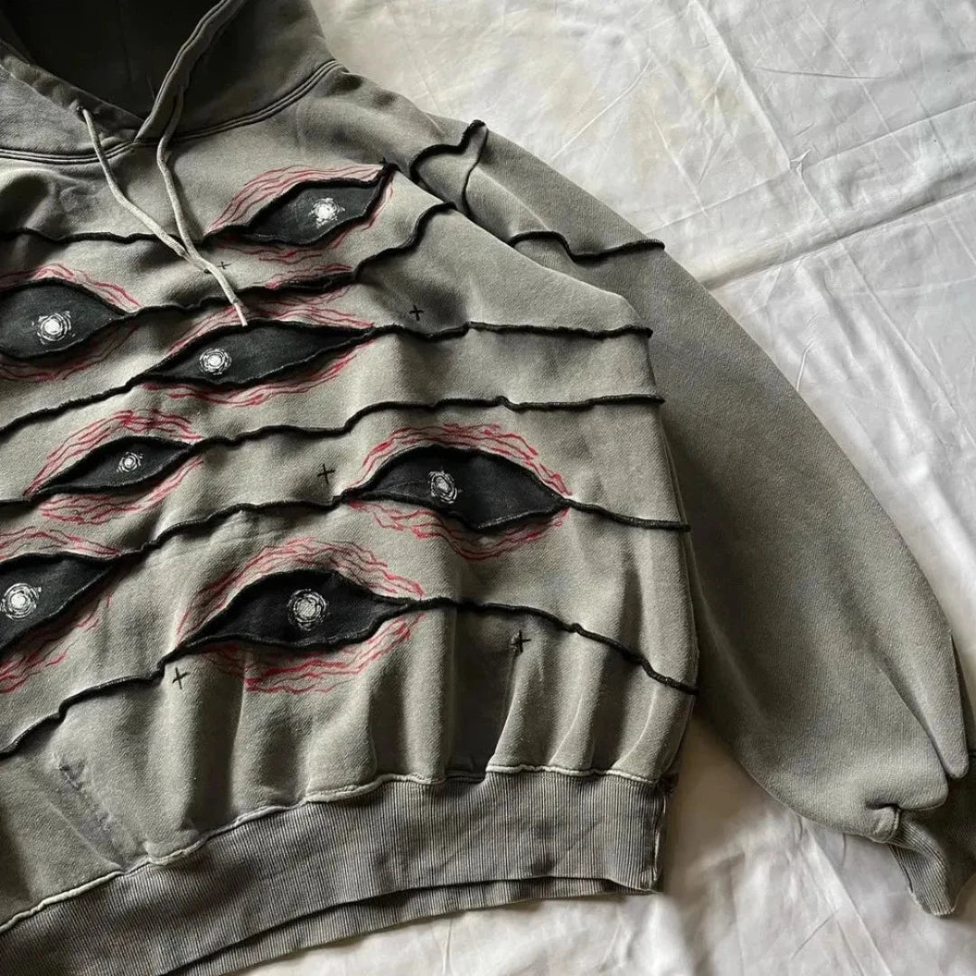 A gray hoodie with an eye pattern patchwork, featuring black and red detailing around the eyes and white centers, displayed on a white sheet.