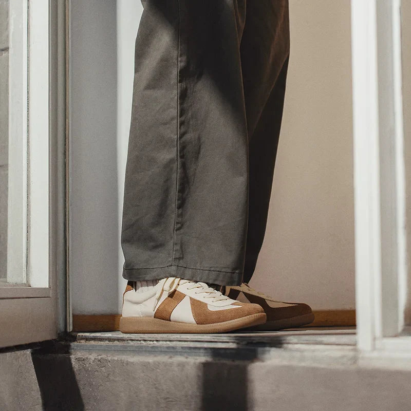 Person standing in a doorway wearing grayish-green pants and tan-and-white lightweight breathable sneakers, with only their lower legs visible. Warm lighting highlights the shoes, which feature EVA midsole cushioning and the bottom of the door frame.