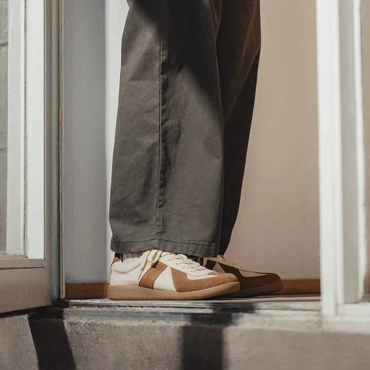 Person standing in a doorway wearing grayish-green pants and tan-and-white lightweight breathable sneakers, with only their lower legs visible. Warm lighting highlights the shoes, which feature EVA midsole cushioning and the bottom of the door frame.