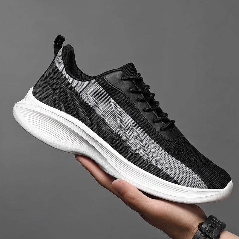 A hand holds up a black and gray athletic sneaker with a white textured sole against a plain gray background. The workout shoe features breathable mesh, black laces, and a pull tab at the heel.