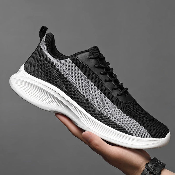 A hand holds up a black and gray athletic sneaker with a white textured sole against a plain gray background. The workout shoe features breathable mesh, black laces, and a pull tab at the heel.