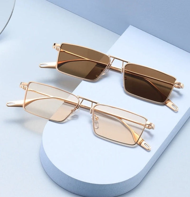 Two pairs of Avalon Sunglasses with rectangular lenses and gold frames, one featuring brown tinted lenses and the other with lightly tinted lenses, displayed on a curved white platform against a light blue background.