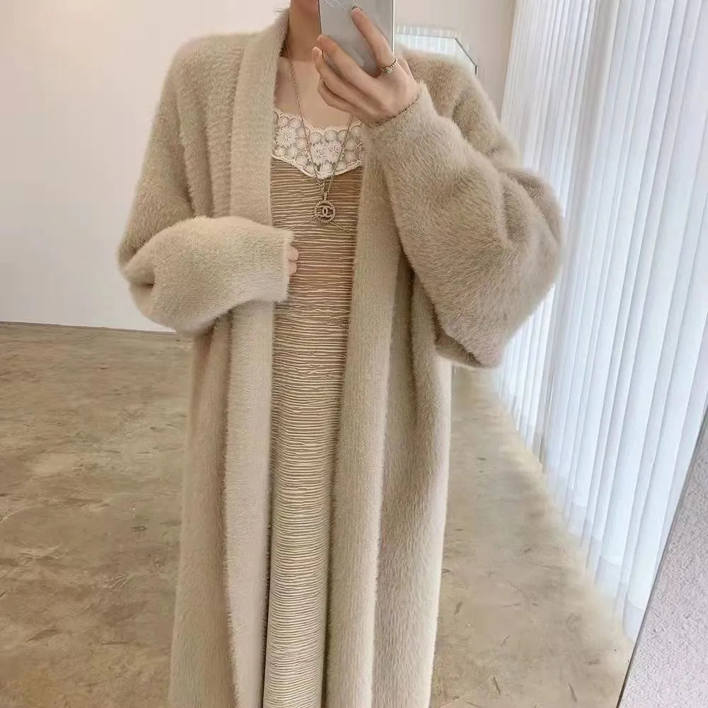 A person stands indoors wearing a long, cashmere blend beige cardigan over a beige dress with lace detail at the neckline, holding a phone to take a mirror selfie. The relaxed drape fit adds elegance against the backdrop of white curtains and a plain wall.
