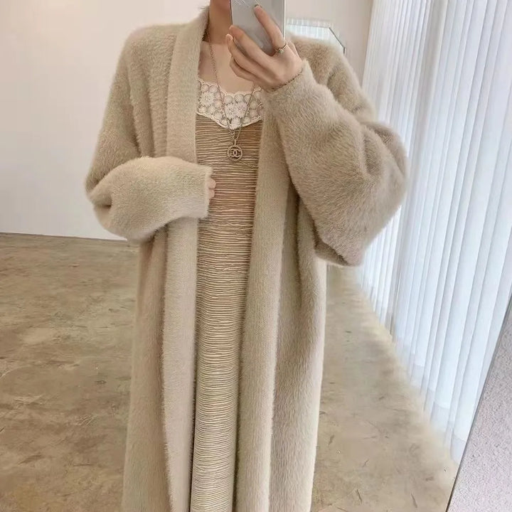 A person stands indoors wearing a long, cashmere blend beige cardigan over a beige dress with lace detail at the neckline, holding a phone to take a mirror selfie. The relaxed drape fit adds elegance against the backdrop of white curtains and a plain wall.