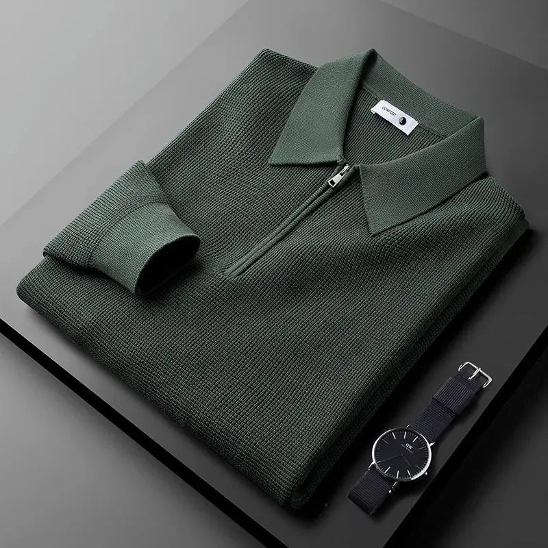 A folded dark green zip-collar long sleeve polo in textured, stretchy fabric lies on a dark surface next to a minimalist black wristwatch with a fabric strap.