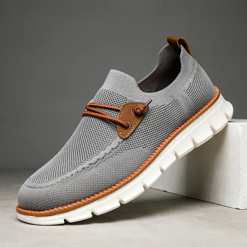 A gray knit lightweight loafer with brown leather accents, white chunky sole, and decorative brown laces, displayed against a plain background with the toe resting on a white block.