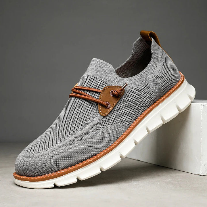 A gray knit lightweight loafer with brown leather accents, white chunky sole, and decorative brown laces, displayed against a plain background with the toe resting on a white block.