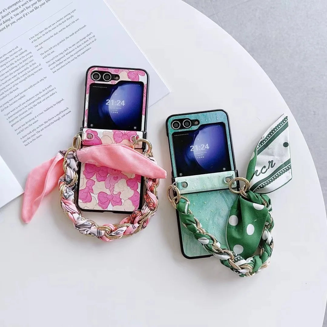 Two smartphones with slim design cases rest on a table. One is pink with flowers and a pink scarf, the other is green with polka dots and a green scarf. Both feature braided chain straps and shockproof protection, placed near an open book.