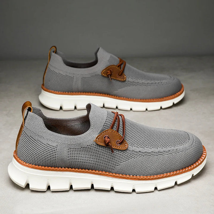 A pair of gray airmesh loafers with white chunky soles, brown leather accents, and decorative brown laces, displayed on a light surface against a gray background.