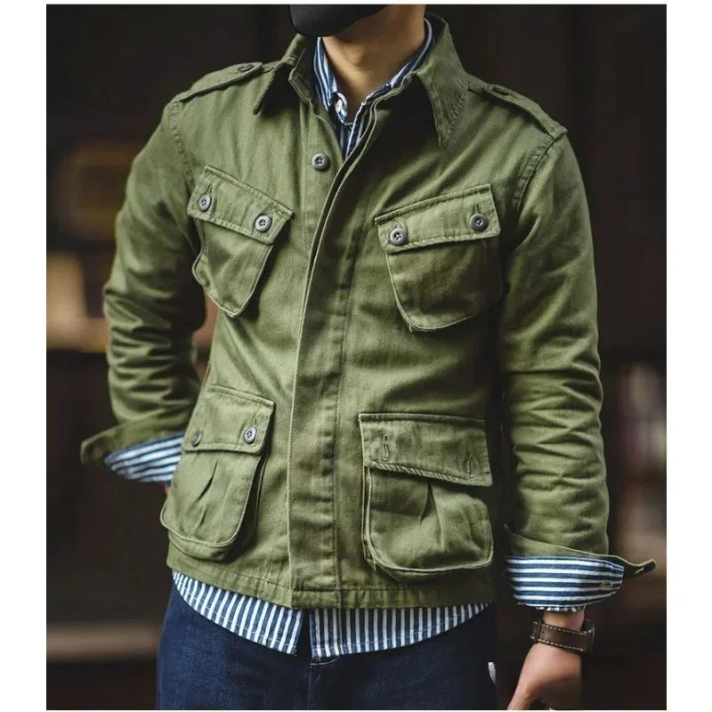 A person wearing a green vintage military jacket with four front pockets over a blue and white striped shirt and dark jeans, standing with hands in pockets. The face is not visible.