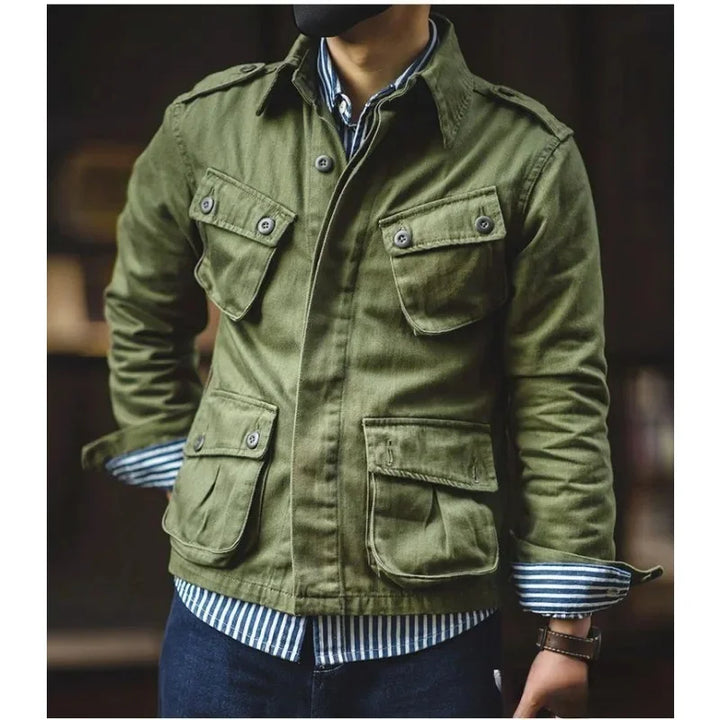 A person wearing a green vintage military jacket with four front pockets over a blue and white striped shirt and dark jeans, standing with hands in pockets. The face is not visible.