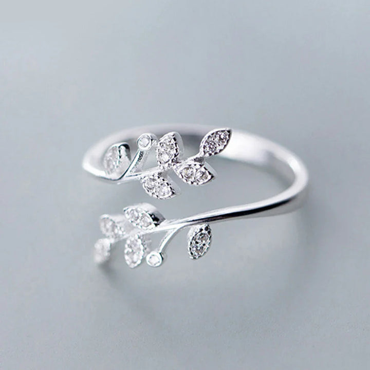 A silver Isadora Floral Ring with an open design features two branches adorned with small, sparkling leaf-shaped gems, displayed on a light gray background—a stunning piece of fine jewelry.
