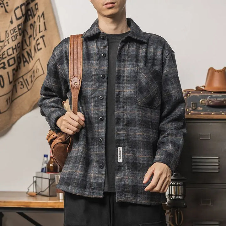 A person wearing a dark plaid oversized shirt and black pants stands indoors with a brown backpack strap over one shoulder, next to a table and metal cabinet with various items in the background, showcasing effortless layering.