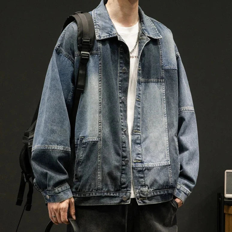 A person wearing an oversized fit, denim patchwork jacket over a white t-shirt, with black jeans and a black backpack, stands against a dark background for a retro style look.