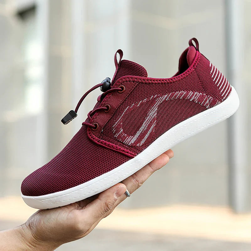 A person holds up a maroon athletic shoe with a white cushioned sole and mesh fabric, featuring a wavy design and white stripes on the heel, showcasing its wide toe box against a blurred outdoor background.