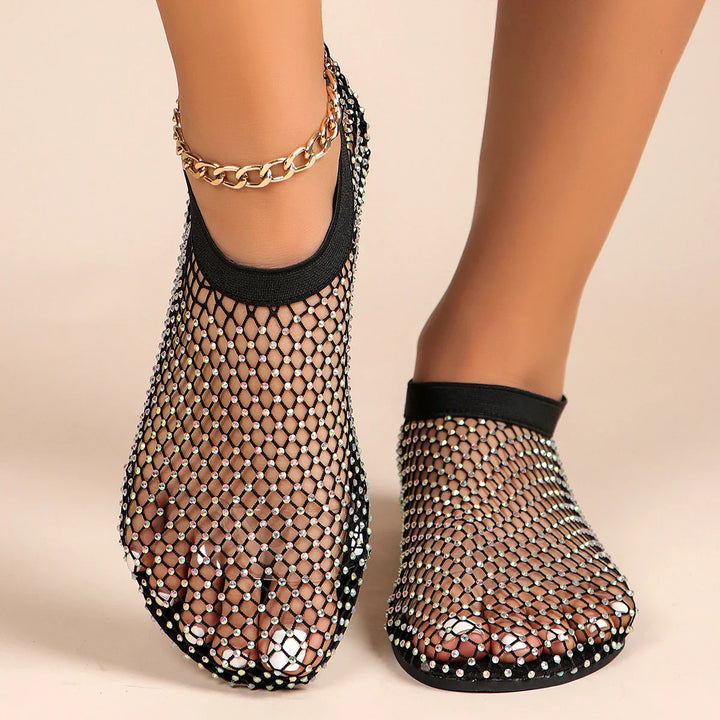 Close-up of a person wearing black mesh overlay shoes with rhinestone accents, revealing toes through the see-through material. One ankle is accessorized with a gold chain bracelet. The background is a neutral beige.