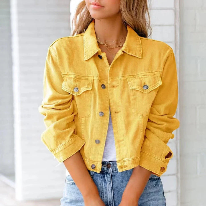 A woman wearing a bright yellow, cropped length denim jacket over a white top and blue jeans, with her hair down, stands against a light-colored brick wall.