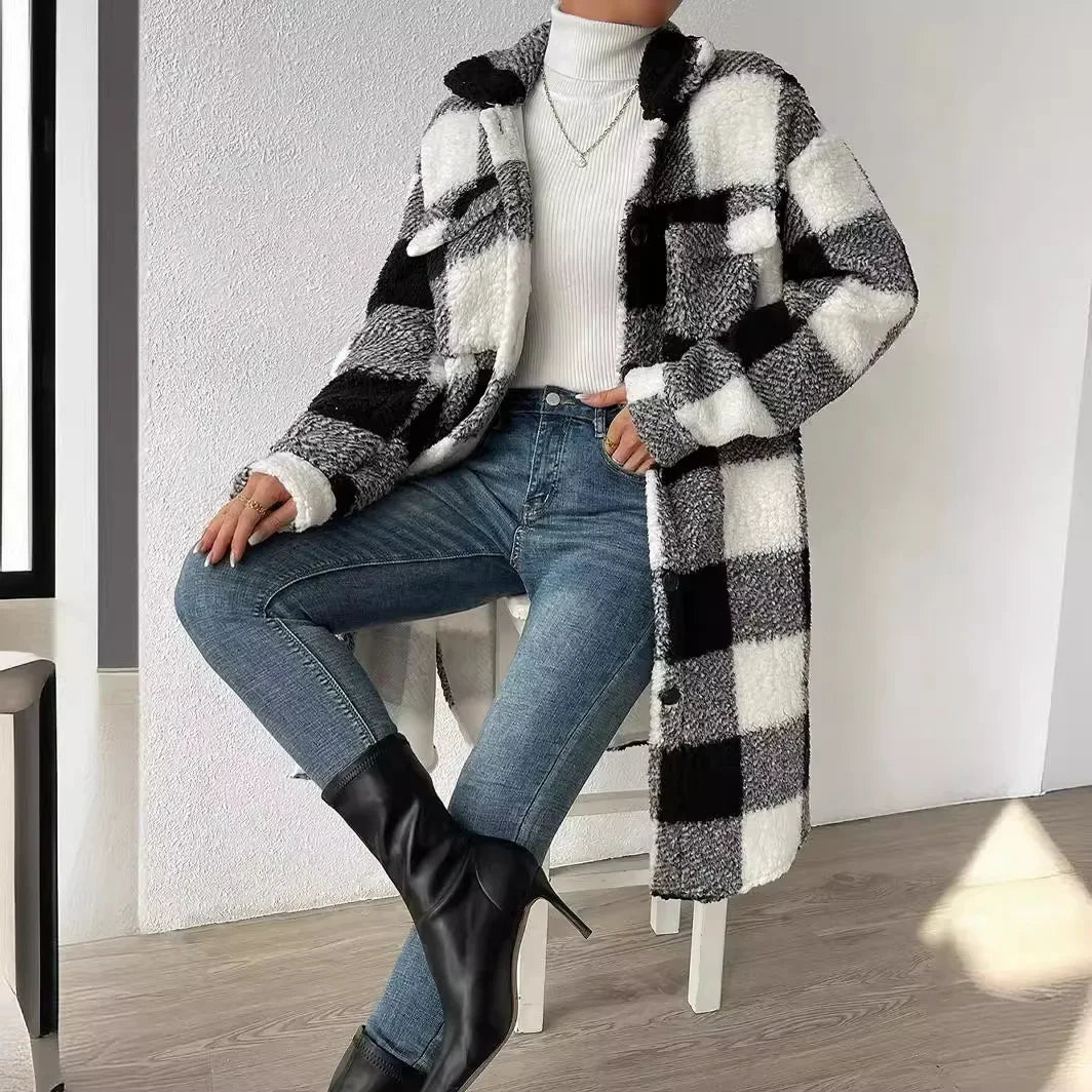 A person wearing a plaid Sherpa open lapel coat, white turtleneck sweater, blue jeans, and black heeled boots sits with legs crossed on a stool in a minimal, sunlit room.
