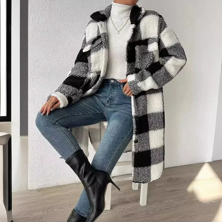 A person wearing a plaid Sherpa open lapel coat, white turtleneck sweater, blue jeans, and black heeled boots sits with legs crossed on a stool in a minimal, sunlit room.