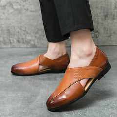 A person wearing stylish, genuine leather loafers with open side cutouts and a durable outsole stands on a gray concrete floor, dressed in black pants.