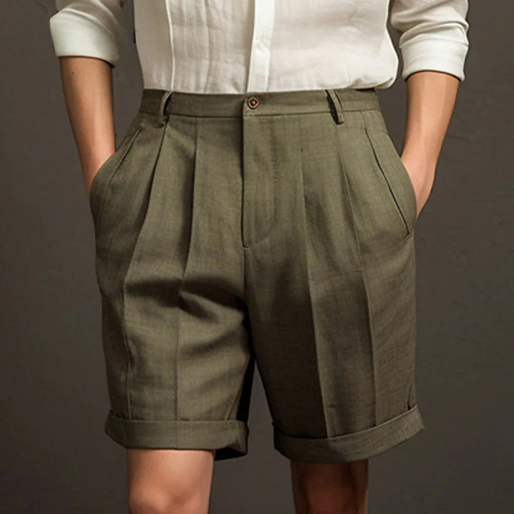A person wears a white long-sleeve shirt and high-waisted, pleated olive green linen shorts with rolled cuffs—perfectly breathable for summer—standing against a plain dark background with hands in the shorts pockets.
