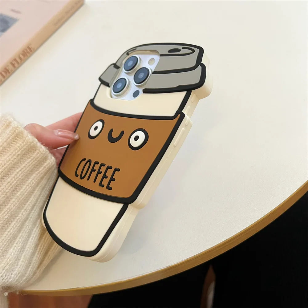 A hand holds a phone with a soft silicone case shaped like a cute 3D coffee cup, featuring a smiling face and the word COFFEE on a brown sleeve. The background shows part of a table and a book, adding charm and shockproof protection.