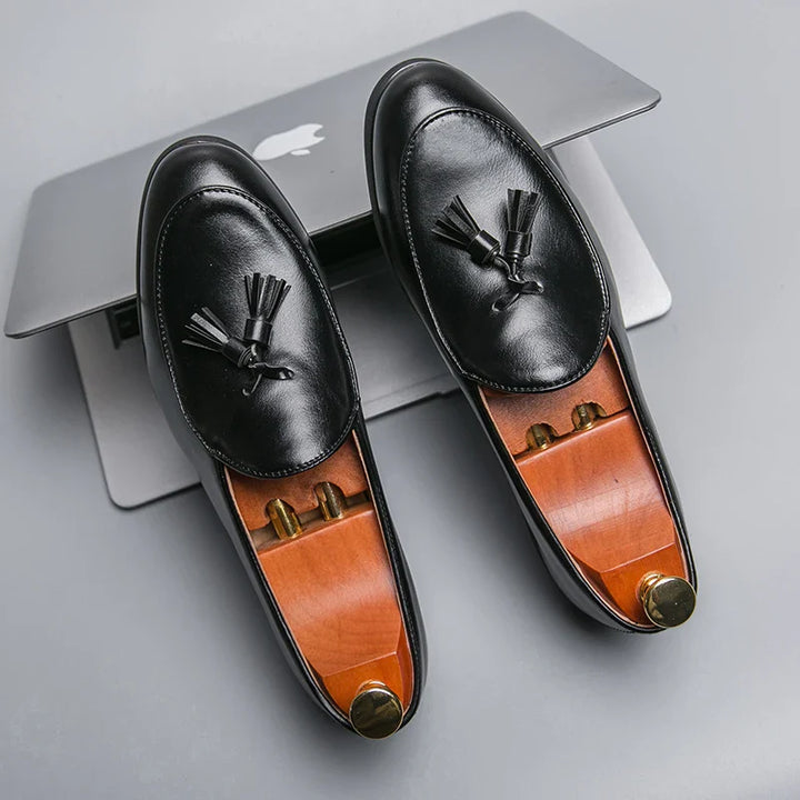 A pair of black leather tassel loafers with a vintage look and wooden shoe trees inside, placed on top of a closed silver laptop on a gray surface.