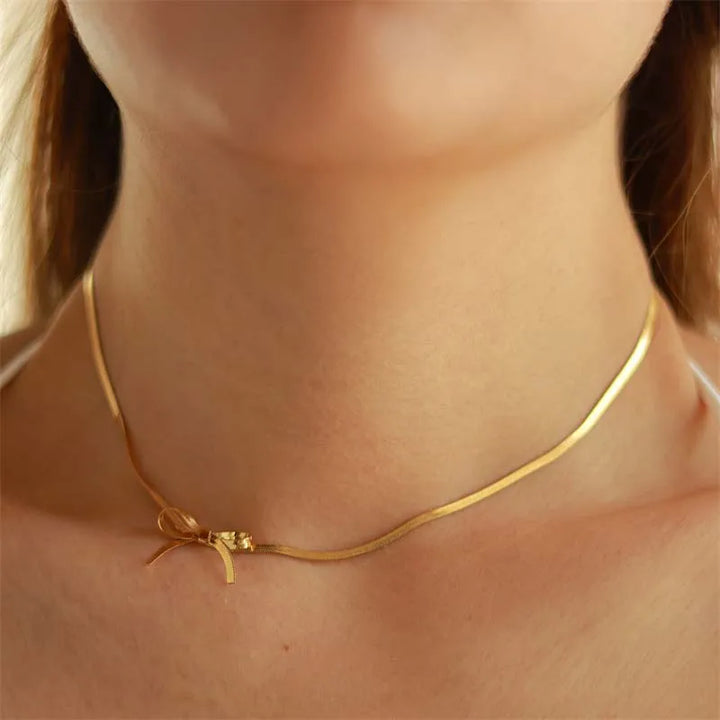 A close-up of a persons neck wearing a thin gold bow necklace with a small detail on the left side. This piece of minimalist jewelry stands out against the softly blurred background.
