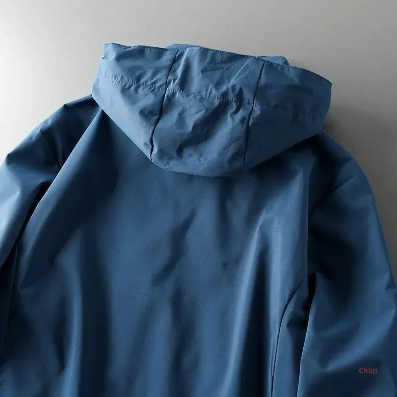 A blue windproof jacket is laid flat on a light gray surface, showing the back and hood. The material appears smooth and slightly matte.