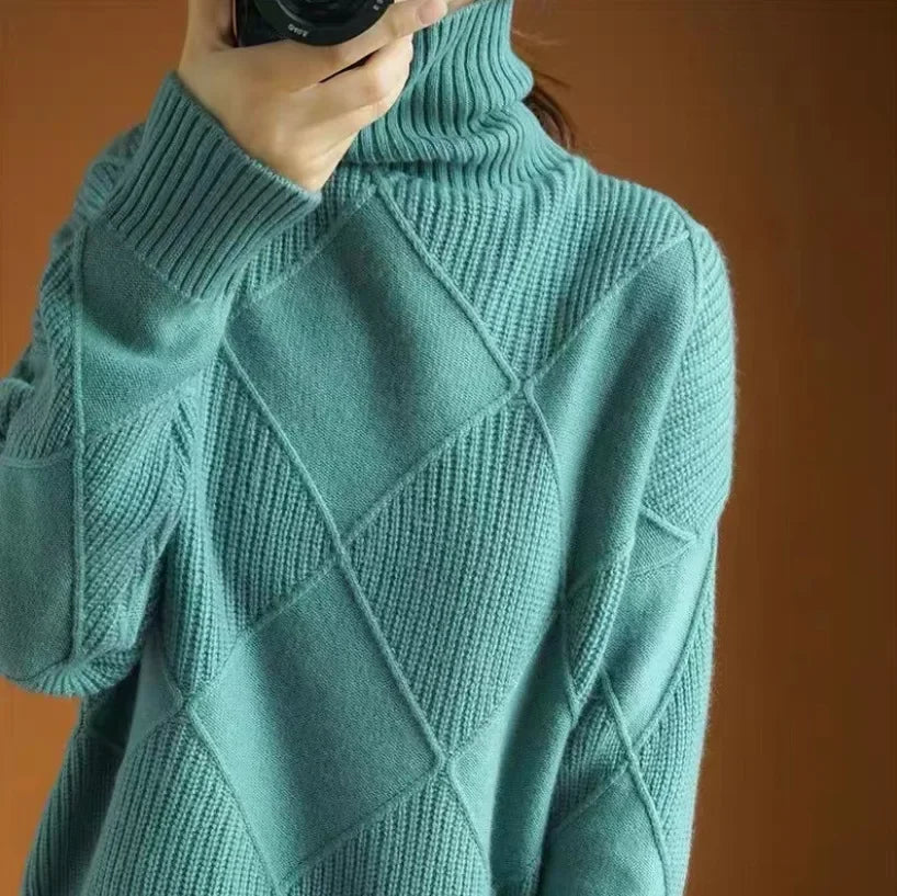 A person wearing a teal, diamond knit sweater with a high neck holds a camera up to their face, obscuring their features. The background is a warm brown color.