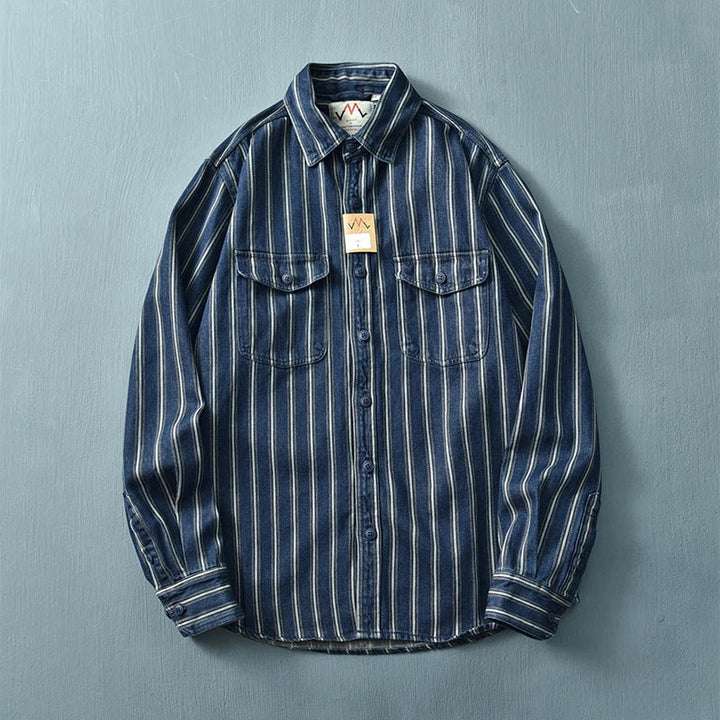 A relaxed fit, long-sleeve denim shirt crafted from heavyweight cotton with vertical white and blue stripes, vintage button-up front, and two chest pockets with flaps, displayed flat against a light blue background.