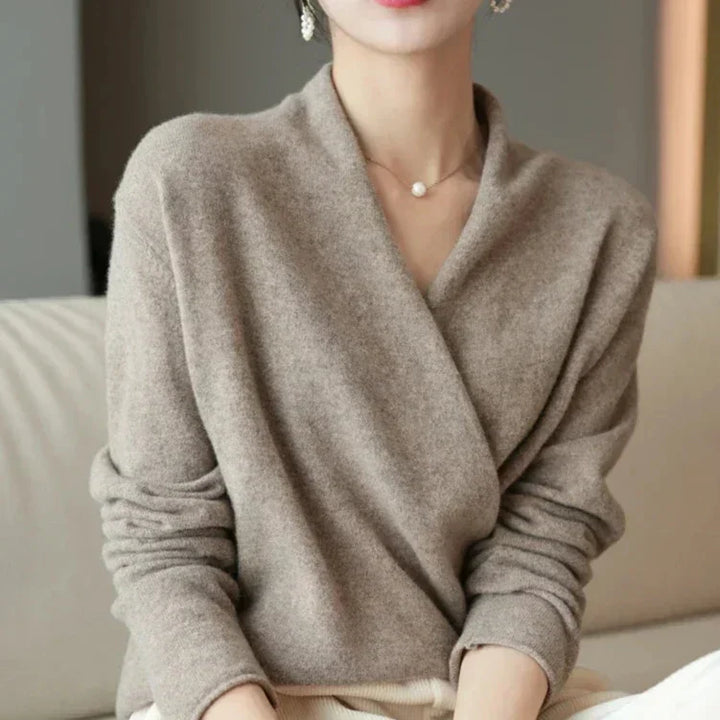 A woman sits on a light-colored sofa, wearing a beige, long-sleeved, wrap-style knit sweater crafted from luxurious cashmere and a delicate necklace. Her face is partially out of frame.