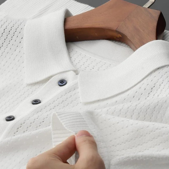 A close-up of a High-End Knitted Polo Shirt featuring a textured, perforated pattern, black buttons, and a ribbed collar—its breathable fabric is highlighted as it's displayed on a wooden hanger, with a hand holding the edge.