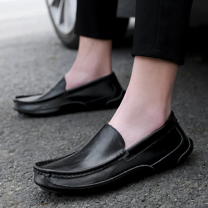 A person wearing genuine leather loafers with a cushioned insole stands on a paved surface near a car tire, dressed in black pants with ankles visible.