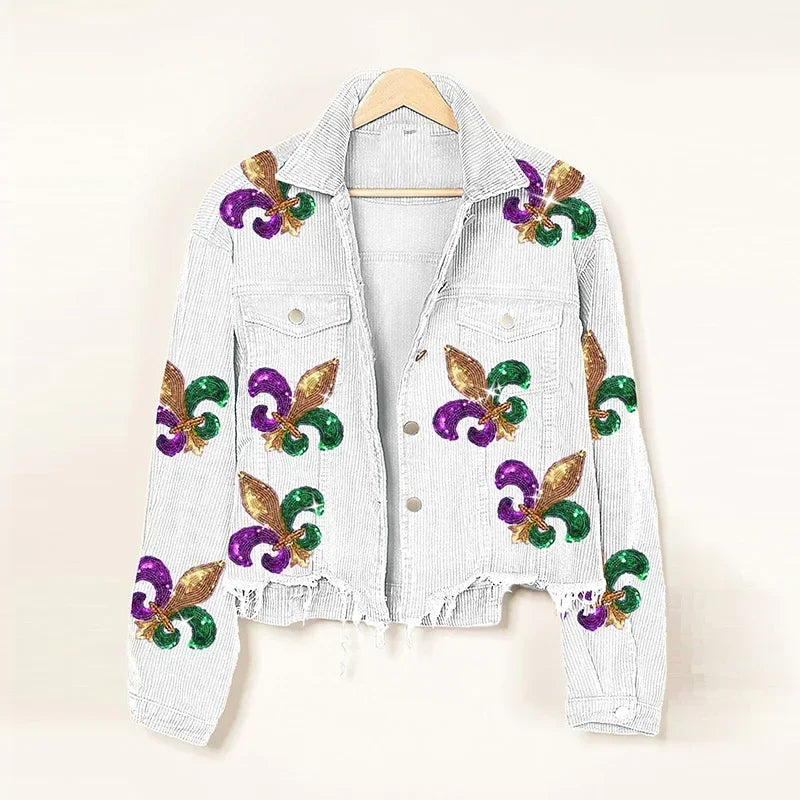 A white denim jacket with frayed edges and a cropped relaxed fit, adorned with glittery gold, green, and purple fleur-de-lis designs, hangs on a wooden hanger against a plain background.