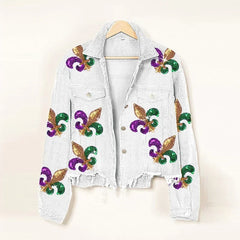 A white denim jacket with frayed edges and a cropped relaxed fit, adorned with glittery gold, green, and purple fleur-de-lis designs, hangs on a wooden hanger against a plain background.